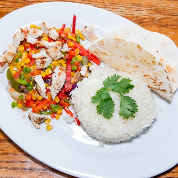 Haitian chicken fajitas with colorful bell peppers, corn, white rice, and tortillas on a white plate.