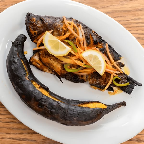 Haitian grilled fish with onions, peppers, lemon slices, and a charred plantain on a white plate.